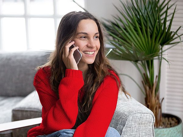 Eine Frau sitzt in ihrem Wohnzimmer auf dem Sofa und telefoniert lächelnd. 