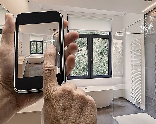 Ein Mann hält ein Smartphone in der Hand, worin der Videomodus aktiviert ist. Dabei filmt er einen Schaden im Badezimmer.