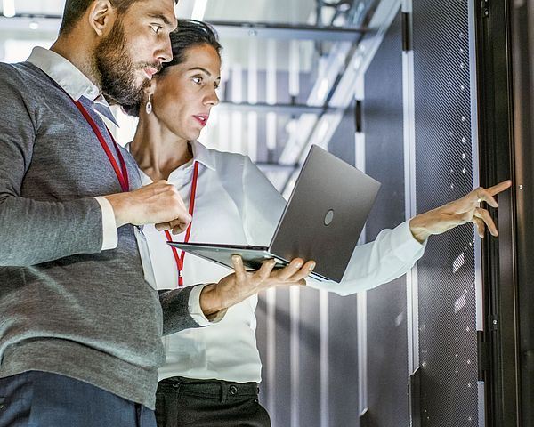 Ein Mann und eine Frau der IT Abteilung stehen mit einem Laptop in einem Rechenzentrum und deuten dabei auf einen Server.