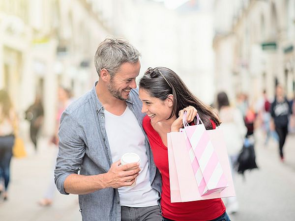 Ein Paar spaziert eine Fußgängerzone entlang. Dabei sehen sie sich lachend an und die Frau hält Einkaufstaschen in der Hand.  