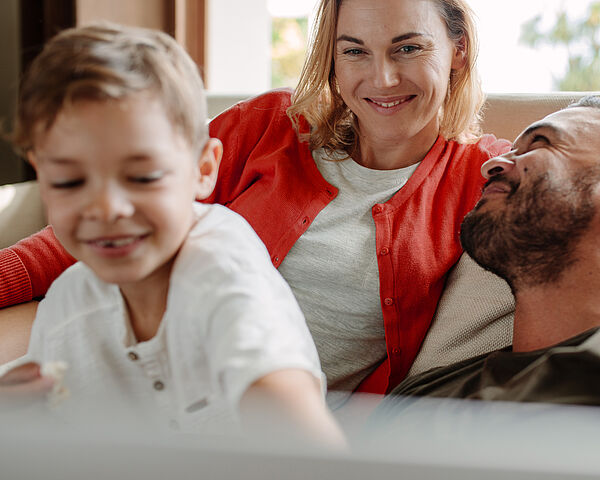 Familiäre Szene im Wohnzimmer: Eine Frau und ein Mann sitzen entspannt auf einem Sofa und lächeln sich an, während ein kleines Kind im Vordergrund spielt und lachend in Bewegung ist.