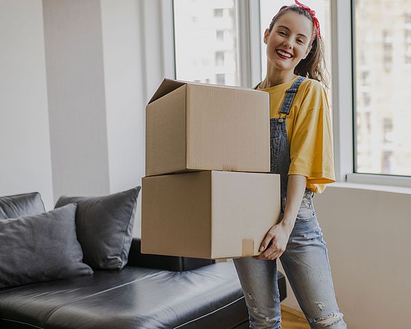 Mädchen steht lächelnd mit zwei Umzugskartons in der Hand in der ersten eigenen Wohnung.