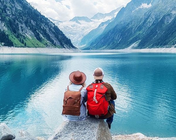 Reisendes Paar sitzt auf einem Felsen und blickt auf einen See in den Bergen.