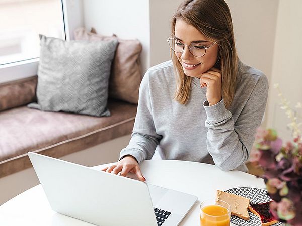 Eine junge Frau arbeitet zu Hause am Laptop während sie frühstückt.