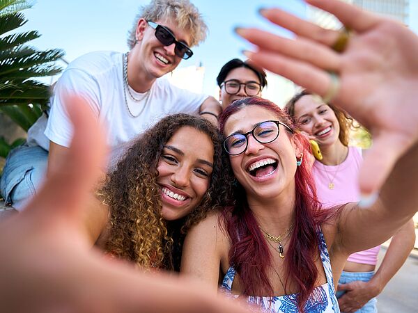 Fünf junge Erwachsene lachen bei einem gemeinsamen Selfie im Freien. Die Stimmung ist fröhlich und ungezwungen.