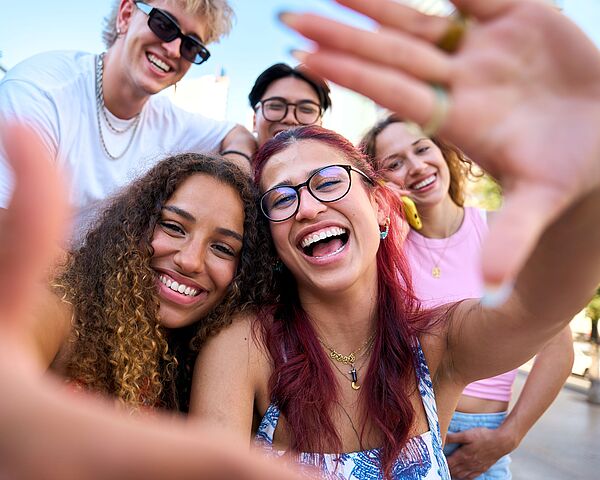 Fünf junge Erwachsene lachen bei einem gemeinsamen Selfie im Freien. Die Stimmung ist fröhlich und ungezwungen.