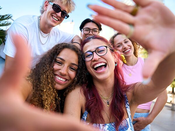 Fünf junge Erwachsene lachen bei einem gemeinsamen Selfie im Freien. Die Stimmung ist fröhlich und ungezwungen.
