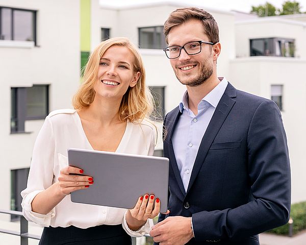 Eine Geschäftsfrau und ein Geschäftsmann befinden sich auf einer Dachterrasse. Dabei lächeln beide in die Kamera und halten ein Tablet in der Hand. 