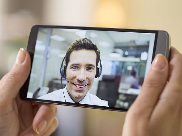 Eine Frau hält ein Smartphone in der Hand, worüber eine Videokonferenz mit einem Berater stattfindet  