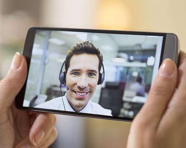 Eine Frau hält ein Smartphone in der Hand, worüber eine Videokonferenz mit einem Berater stattfindet  