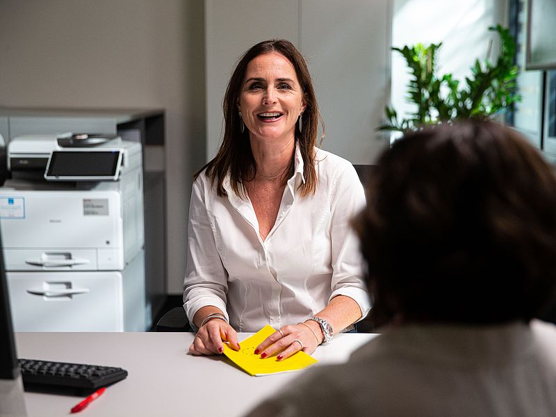 Dunkelhaarige Frau sitzt am Schreibtisch und lacht.