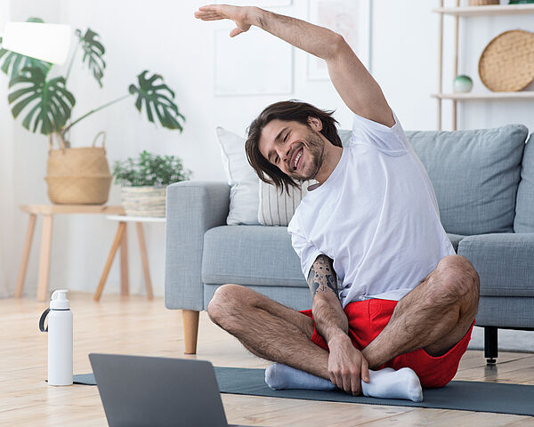 Ein junger Mann macht Gymnastikübungen zu Hause auf seiner Yoga-Matte vor seinem Laptop