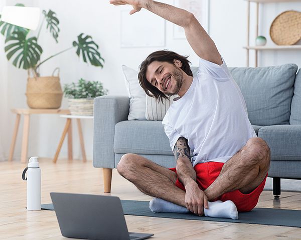 Ein junger Mann macht Gymnastikübungen zu Hause auf seiner Yoga-Matte vor seinem Laptop