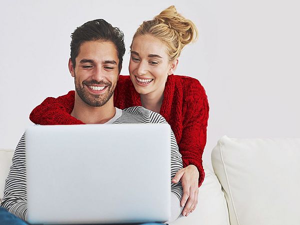 Ein junges glückliches Paar sitzt auf der Couch und blickt auf einen Laptop. Die Frau umarmt den Mann dabei von hinten.