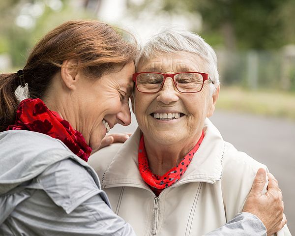 Zwei ältere Frauen befinden sich auf einem Spaziergang und umarmen sich lächelnd 