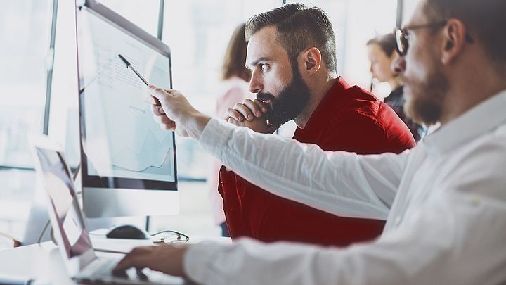 Zwei Männer befinden sich in einem Büro am Schreibtisch und begutachten dabei einen Bericht auf einem Bildschirm  