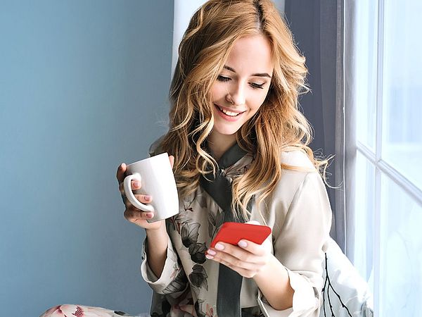 Eine junge Frau sitzt am Fensterbrett mit einer Tasse in der Hand und blickt fröhlich auf ihr Smartphone.