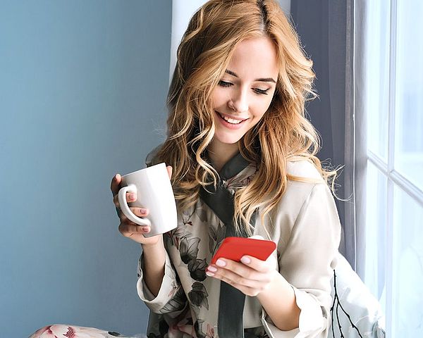 Eine junge Frau sitzt am Fensterbrett mit einer Tasse in der Hand und blickt fröhlich auf ihr Smartphone.