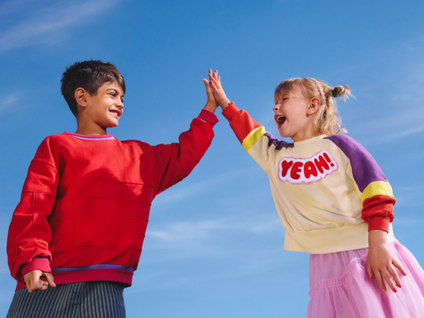 Zwei lachende Kinder geben sich vor blauem Himmel ein High Five. Ein Kind trägt einen bunten Pullover mit der Aufschrift ‚YEAH!‘, das andere einen roten Pullover.
