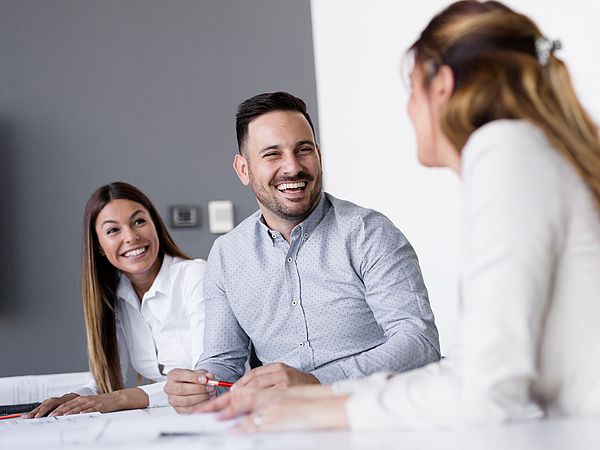 Zwei junge Frauen und ein junger Mann sitzen am Besprechungstisch und lachen.