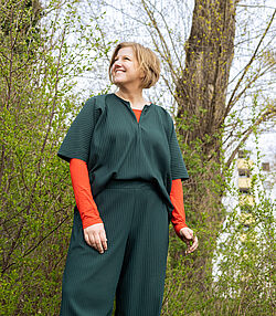 Lächelnde Frau mit kurzem blondem Haar im Freien, die ein dunkelgrünes Outfit mit einem orangen Langarmshirt darunter vor Bäumen und grünem Gebüsch trägt.