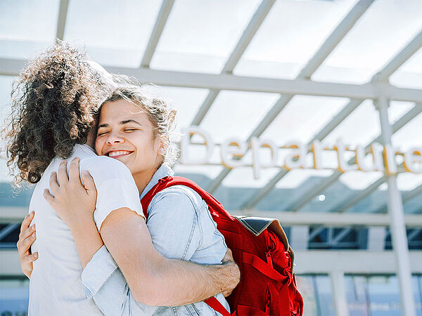 Ein Mann mit langen, gelockten, dunklen Haaren und eine Frau mit dunklen Haaren und rotem Rucksack umarmen sich