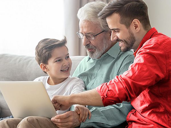 Drei Männer unterschiedlicher Generationen sitzen gemeinsam vor einem Laptop und lächeln sich an.
