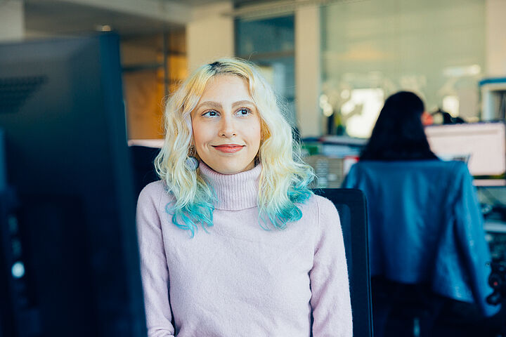 In einem Büro sitzt eine junge Frau mit schulterlangen, blond-türkisen Haaren. Sie trägt einen rosa Rollkragenpullover, blickt auf die Seite und lächelt. Im Hintergrund sieht man eine andere Frau an einem Schreibtisch sitzen, eine blaue Jacke über die Stuhllehne gehängt.
