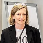 Eine Frau mit hellen Haaren trägt ein weißes Shirt mit Aufdruck und ein schwarzes Sakko. Sie trägt die Brille im Haar und steht vor einem Flipchart.