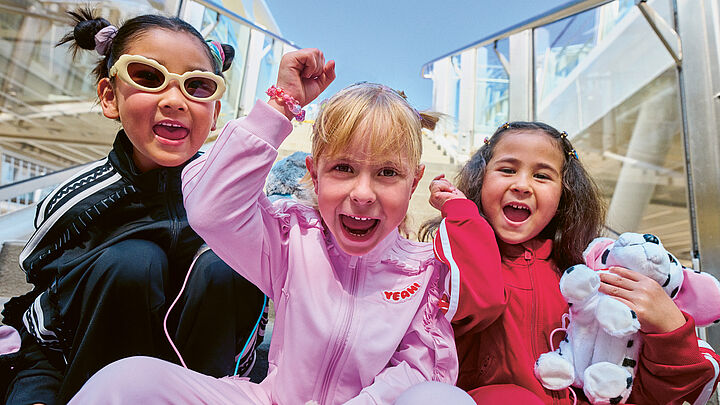 Drei Kinder in farbenfroher Sportkleidung sitzen dicht beieinander und jubeln in die Kamera.