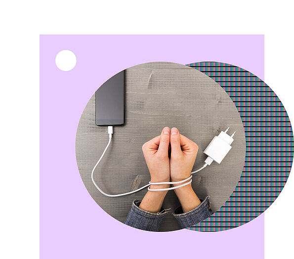 Zwei zur Faust geformte Hände liegen auf einer grauen Tischplatte, die Handgelenke sind mit einem weißen Ladekabel gefesselt, das an einem Smartphone hängt.