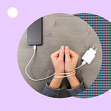 Zwei zur Faust geformte Hände liegen auf einer grauen Tischplatte, die Handgelenke sind mit einem weißen Ladekabel gefesselt, das an einem Smartphone hängt.