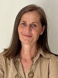 Eine Frau mit schulterlangen braunen Haaren trägt eine beige Bluse und eine lange Kette. Sie neigt den Kopf etwas zur Seite und lächelt.