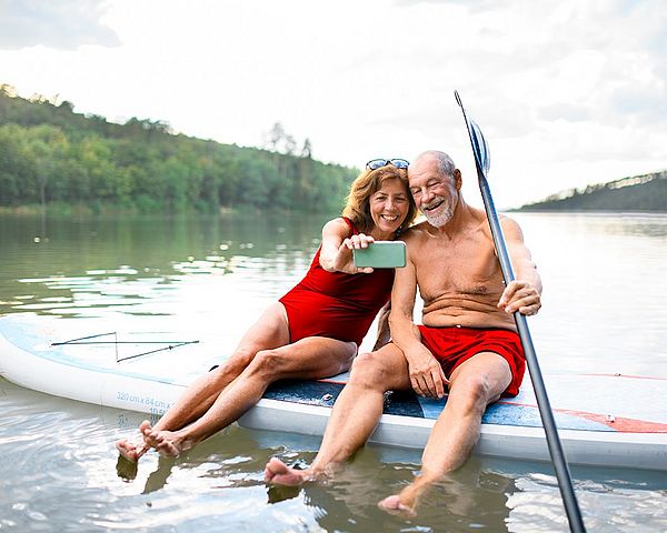 Ein Seniorenpaar sitzt auf einem Paddleboard am See und macht ein Selfie.