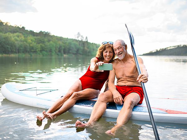 Ein Seniorenpaar sitzt auf einem Paddleboard am See und macht ein Selfie.