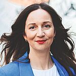 Frau mit langen dunklen Haaren mit blauem Blazer und weißem Shirt steht vor einer Graffittiwand und lächelt.