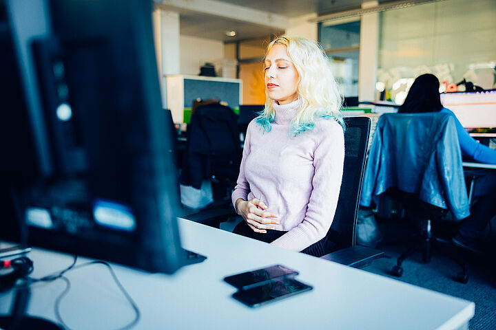 Eine junge Frau mit schulterlangen blond-türkisen Haaren sitzt am Schreibtisch. Ihre Augen sind geschlossen, eine Hand liegt auf ihrem Bauch.