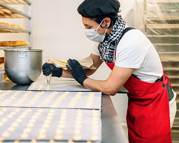 Ein Konditormeister steht in seiner Backstube mit Maske und Handschuhen. Dabei formt er voller Konzentration diverse Gepäcksorten. 