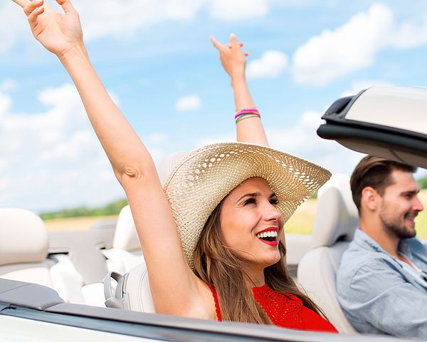 Ein junges glückliches Paar fährt gemeinsam in einem Cabrio. Die Frau streckt vor Freude ihre Arme in die Luft.