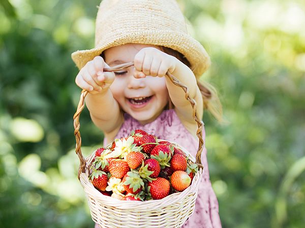 Ein junges Mädchen mit Sonnenhut im Kleid, befindet sich auf einer Wiese und streckt voller Freude einen Korb mit Erdbeeren in die Höhe. 