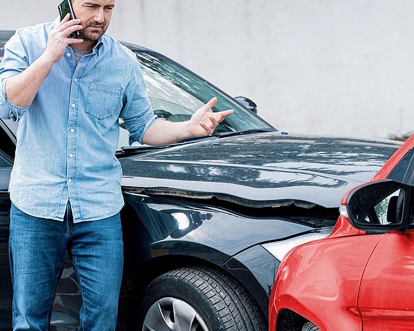 Ein Mann telefoniert voller Aufregung nach einem Autounfall. Im Hintergrund sind dabei zwei Autos zu sehen, welche frontal zusammengestoßen sind. 