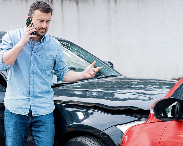 Ein Mann telefoniert voller Aufregung nach einem Autounfall. Im Hintergrund sind dabei zwei Autos zu sehen, welche frontal zusammengestoßen sind. 