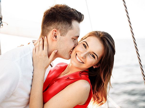 Ein glückliches junges Paar entspannt auf einem Boot am Meer. Der Mann gibt der Frau einen Kuss auf die Wange.