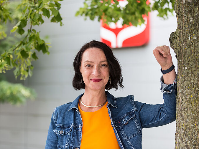 Frau steht an Baum gelehnt und lächelt in die Kamera.