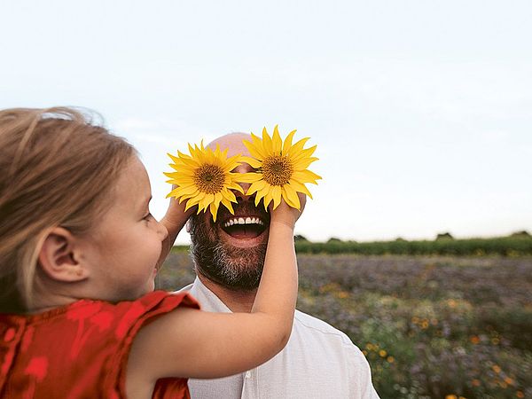 Ein junges Mädchen hält ihrem Vater zwei Sonnenblumen vor die Augen. Beide befinden sich auf einer Blumenwiese. 