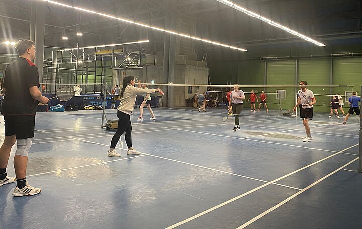 Dynamische Szene eines Badminton-Turniers in einer Turnhalle. Im Vordergrund spielen zwei Teams im Doppel gegeneinander über das Netz.