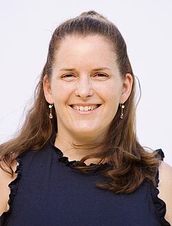 Porträtfoto einer Frau mit schulterlangen braunen Haaren, in einem dunkelblauen ärmellosen Shirt, mit Hängeohrringen.