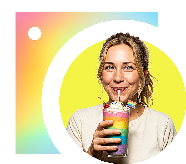 Eine lächelnde Frau mit Sommersprossen und zusammengebundenen Haaren trinkt aus einem geschichteten Regenbogen-Milchshake, der mit Schlagsahne, einer Kirsche und einer kleinen Pride-Flagge dekoriert ist, vor einem runden, hellgelben Hintergrund.