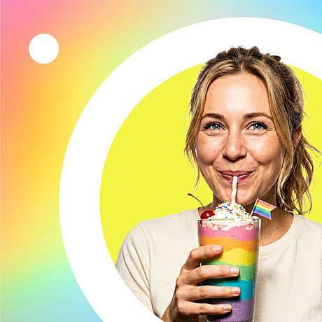 Eine lächelnde Frau mit Sommersprossen und zusammengebundenen Haaren trinkt aus einem geschichteten Regenbogen-Milchshake, der mit Schlagsahne, einer Kirsche und einer kleinen Pride-Flagge dekoriert ist, vor einem runden, hellgelben Hintergrund.