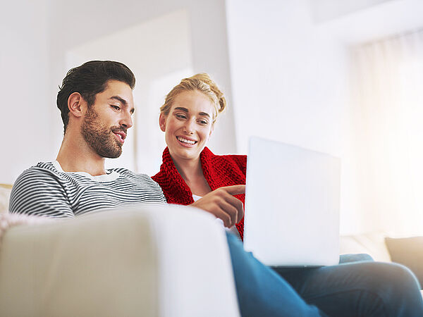 Ein junger Mann sitzt mit seiner Freundin auf der Couch und zeigt ihr etwas auf seinem Laptop.